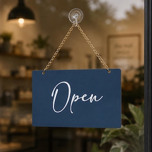 May include: A navy blue rectangular sign with the word "Open" in white script lettering hangs from a gold chain. The sign is attached to a glass door with a suction cup. The background is blurred, showing the interior of a shop.