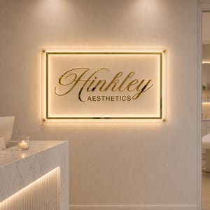 Può includere: Un'insegna rettangolare con il nome "Hinkley Aesthetics" in caratteri dorati. L'insegna è illuminata da una luce calda e incorniciata in oro. È montata su una parete di colore neutro.