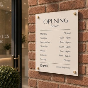 May include: A cream-coloured rectangular sign displaying business opening hours, mounted on a red brick wall. The sign features the words "OPENING hours" in dark grey, with a list of opening times for each day of the week and social media icons.