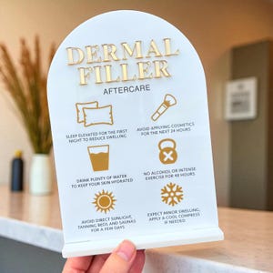 May include: A white acrylic sign with gold lettering that reads "DERMAL FILLER AFTERCARE." The sign includes aftercare instructions with icons and text, such as avoiding cosmetics and direct sunlight. The sign is held by a hand.
