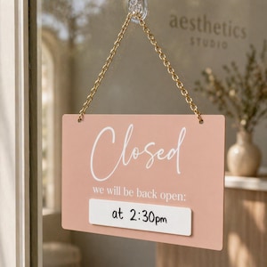 Puede incluir: Un letrero rectangular de color melocotón con la palabra "Closed" en escritura blanca. Debajo, se lee "we will be back open: at 2:30pm" en una fuente más pequeña. El letrero cuelga de una cadena dorada.