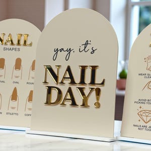 May include: Cream-coloured sign with gold lettering that reads "yay, it's NAIL DAY!". The sign is part of a set, with other signs displaying nail shapes and nail care tips. The signs have a rounded top and sit on a white base.