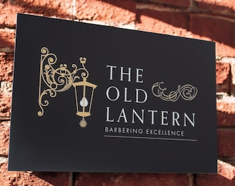 May include: A black sign with gold lettering that reads "The Old Lantern Barbering Excellence". The sign features a stylized image of a lantern.