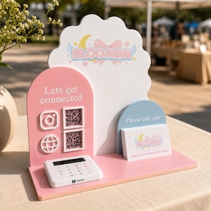 Puede incluir: Un expositor rosa y blanco con el texto "BLOOMIMI" y "Lets get connected". Incluye un terminal de pago, iconos de redes sociales, códigos QR y tarjetas de visita.