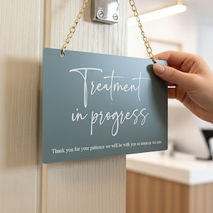 May include: A rectangular sign in a muted teal colour, with the words "Treatment in progress" in white script. It hangs from a gold chain. The text "Thank you for your patience we will be with you as soon as we can" is printed at the bottom of the sign.