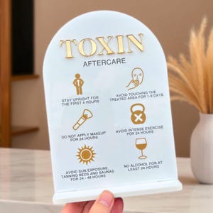 May include: A white acrylic sign with gold lettering that reads "TOXIN AFTERCARE." The sign includes illustrated aftercare instructions: stay upright, avoid touching the area, do not apply makeup, avoid intense exercise, avoid sun exposure, and no alcohol.