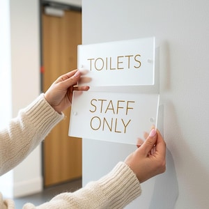 May include: Two rectangular frosted acrylic signs with gold lettering. The top sign reads "TOILETS", and the bottom sign reads "STAFF ONLY". The signs are being held up against a white wall, ready to be mounted.