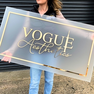 May include: A frosted glass sign with a gold frame and gold lettering that reads "VOGUE Aesthetics".
