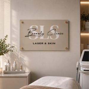 Op de afbeelding: Een beige bord met gouden accenten met de tekst "SLS Beauty Clinic Laser & Skin" in zwarte en witte letters. Het bord is bevestigd aan een lichtbeige muur. Een behandelbed staat op de voorgrond.