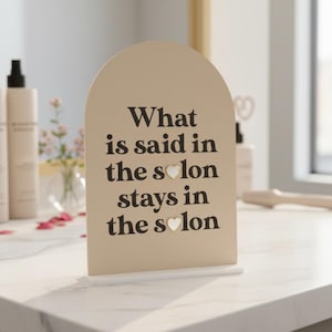 Könnte beinhalten: Beiges, bogenförmiges Schild mit dem Text "What is said in the salon stays in the salon" in Schwarz. Das Wort "Salon" hat ein Herzsymbol anstelle des "o". Das Schild steht auf einem weißen Sockel.
