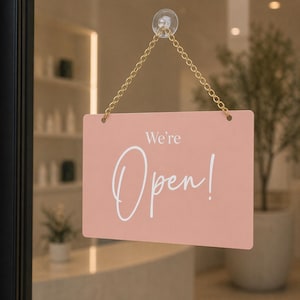 May include: A rectangular, pale pink sign with the words "We're Open!" in white script lettering. The sign is suspended by a gold chain and suction cup, indicating a business is open.