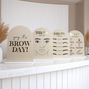 May include: Four beige arched signs with gold lettering and graphics related to eyebrow care. The signs display text such as "Brow Day!", "Brow Mapping", "Brow Shapes", and "Brow Aftercare", with illustrations of eyebrows and aftercare instructions.