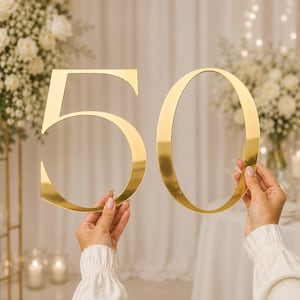 Può includere: Un grande numero 50 ritagliato di colore dorato e riflettente. Il numero cinque e il numero zero sono cifre separate, piatte, con una finitura metallica e una superficie liscia, tenute in mano per mostrare chiaramente i numeri su uno sfondo neutro.