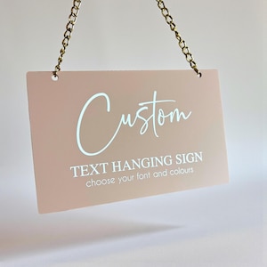 Pode incluir: Uma placa retangular rosa claro pendurada por uma corrente dourada. A placa apresenta a palavra "Custom" em uma fonte cursiva, com o texto "TEXT HANGING SIGN choose your font and colours" abaixo.