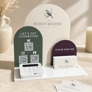 Peut inclure: Un présentoir à plusieurs niveaux comprenant une grande arche crème avec un logo d'oiseau et du texte, une arche vert foncé avec trois codes QR et des icônes de réseaux sociaux, et une petite arche violet foncé avec un porte-cartes de visite.