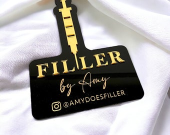 May include: Black acrylic sign with gold lettering that reads "FILLER by Amy" and a gold Instagram icon with the text "@AMYDOESFILLER". The sign features a gold silhouette of a syringe.