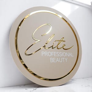 以下が含まれることがあります： 金色の縁取りが付いた丸いベージュの看板に、白い文字で「Elite Professional Beauty」と書かれている。