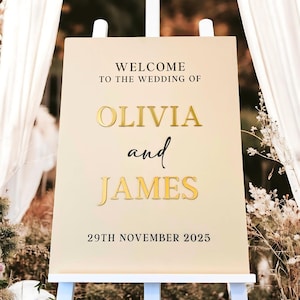 Op de afbeelding: Een beige bruiloft welkom bord met gouden letters. Het bord staat "Welkom op de bruiloft van Olivia en James 29 november 2025".