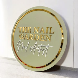 Może przedstawiać: Okrągły szyld z szałwiowym zielonym tłem i złotą obwódką. Tekst "THE NAIL GARDEN" jest w kolorze złotym, a "Nail Artist" w białym skrypcie. Znak jest prawdopodobnie dla salonu paznokci.
