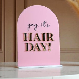 Może przedstawiać: Różowy akrylowy znak z zaokrąglonym wierzchołkiem i białą podstawą. Znak zawiera frazę "yay, it's HAIR DAY!" w czarnym skrypcie i złotych, wypukłych literach. Znak jest przeznaczony dla salonu fryzjerskiego lub firmy kosmetycznej.