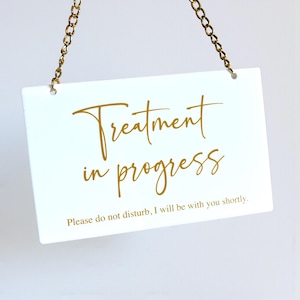 May include: A white rectangular sign with gold text that reads "Treatment in progress." The sign is suspended by a gold chain. The bottom of the sign reads "Please do not disturb, I will be with you shortly."