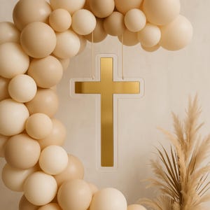 Peut inclure: Une croix dorée sur fond blanc est suspendue à une ficelle contre un mur beige. Une guirlande de ballons beiges se trouve en arrière-plan.