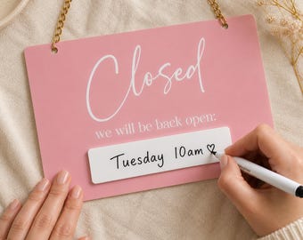 May include: A pink rectangular sign with the word "Closed" in white script. Below, it reads "we will be back open:" and "Tuesday 10 am ♡" is written on a white rectangle. A gold chain hangs from the top.