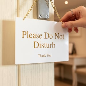 Könnte beinhalten: Ein weißes, rechteckiges Türschild mit goldenem Text, der „Please Do Not Disturb“ und „Thank You“ lautet. Das Schild wird von einer goldenen Kette an einer silbernen Halterung aufgehängt.