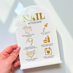 Puede incluir: Un letrero acrílico blanco con letras doradas que dice "NAIL AFTERCARE". El letrero presenta consejos ilustrados para el cuidado de las uñas, incluido el uso de guantes, la aplicación de aceite para cutículas y la hidratación. El letrero lo sostiene una persona.