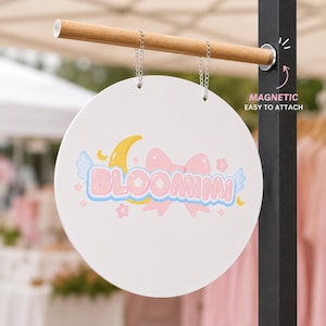 May include: A white, round sign with the word "BLOOMIMI" in pastel colors, featuring a crescent moon and bow design. The sign hangs from a wooden rod with metal chains. Text on the side reads "MAGNETIC EASY TO ATTACH".