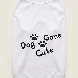 Op de afbeelding: Witte honden-T-shirt met zwarte tekst en pootafdrukken. De tekst luidt "Dog Gone Cute". Het shirt is mouwloos, waarschijnlijk voor een kleine hond of puppy.