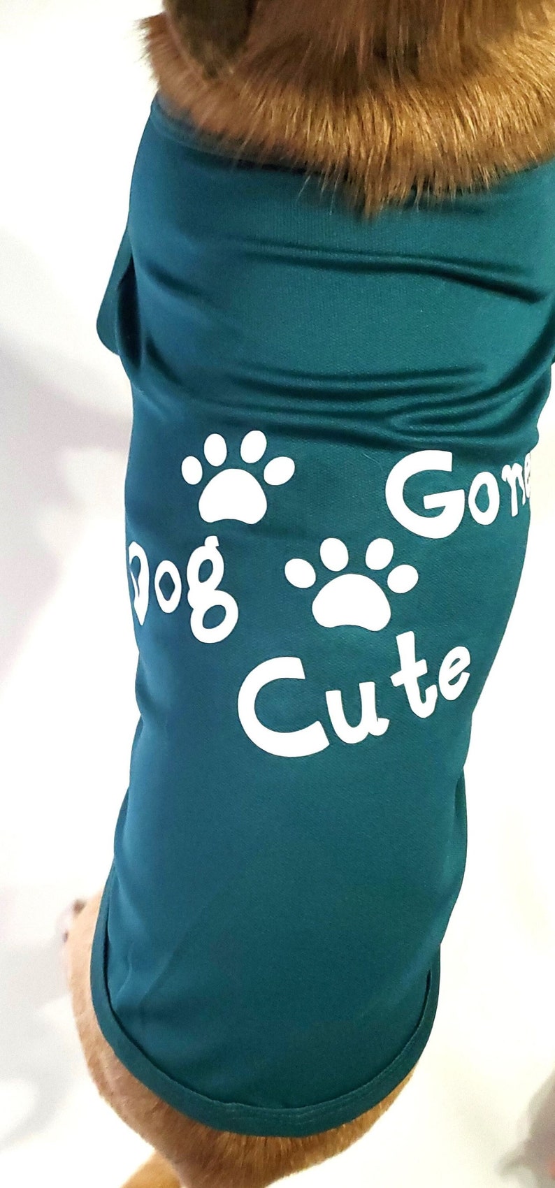 Op de afbeelding: Teal honden-T-shirt met witte pootafdrukken en de woorden "Dog Gone Cute". Het shirt is gemaakt van een zacht materiaal en is ontworpen om een hond te passen. De vacht van de hond is zichtbaar aan de bovenkant.
