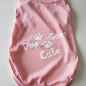 Op de afbeelding: Een lichtroze honden-T-shirt met de tekst "Dog Gone Cute" en witte pootafdrukken. Het shirt heeft een ronde halslijn en is gemaakt van een zachte stof.