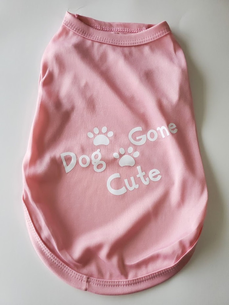 Op de afbeelding: Een lichtroze honden-T-shirt met de tekst "Dog Gone Cute" en witte pootafdrukken. Het shirt heeft een ronde halslijn en is gemaakt van een zachte stof.
