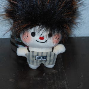 May include: A plush doll with a large tuft of black and brown faux fur hair, a white face with rosy cheeks, black eyes, and a red nose. The doll wears beige and black plaid overalls with the word COOK embroidered on a white patch on the front.