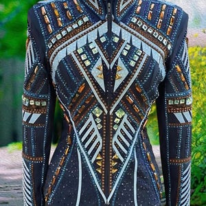 Puede incluir: Chaqueta de concurso ecuestre azul marino con intrincados adornos. La chaqueta presenta un cuello alto y una cremallera completa. Está adornada con patrones geométricos de cristales dorados, marrones y transparentes, y detalles en blanco. Las mangas tienen un diseño a rayas.