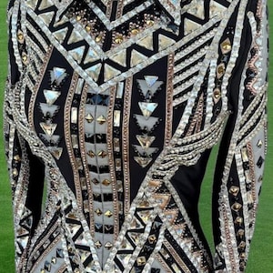 Op de afbeelding: Een zwart showjack met een geometrisch patroon met zilveren en gouden strass steentjes. Het jack heeft een ritssluiting en lange mouwen.