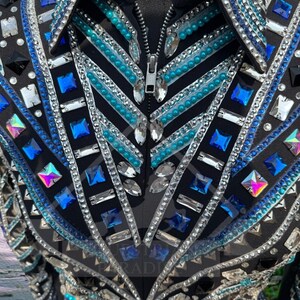 Könnte beinhalten: Schwarze Reitjacke mit aufwendigen Verzierungen. Sie hat einen Reißverschluss, einen Kragen und ist mit einer schillernden Anordnung aus blauen, silbernen und irisierenden Kristallen und Strasssteinen verziert. Die Jacke hat eine taillierte Passform und viele Details.