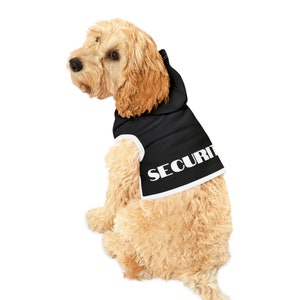 Op de afbeelding: Een lichtbruine hond die een zwarte hoodie-vest draagt met witte bies en het woord "SECURITY" erop gedrukt.