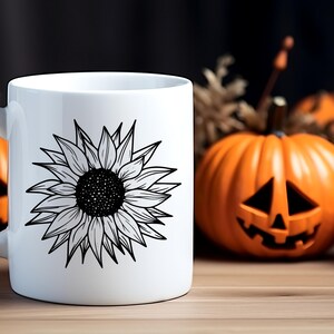 May include: White ceramic mug featuring a detailed black and white sunflower design. The mug is set on a wooden surface with a blurred background that includes an orange jack-o'-lantern and other fall decorations, creating a seasonal theme.