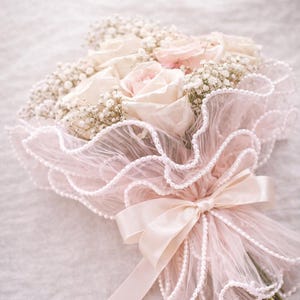 May include: A bouquet of light pink and white roses and baby's breath wrapped in a delicate pink lace with pearl accents. The bouquet is tied with a satin ribbon. Text on the image reads "Luxury Bouquet Wrap."