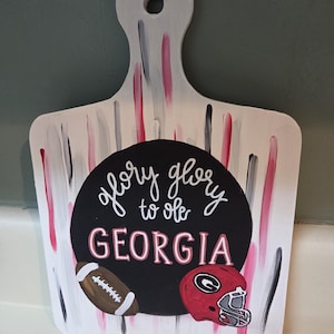 May include: White wooden cutting board with a handle, decorated with red, black, and gray brushstrokes. A black circle features the text "glory glory to ole GEORGIA", a football, and a red football helmet.