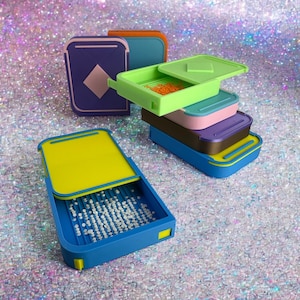 May include: A collection of colorful, rectangular craft organizers with hinged lids. The organizers are in various colors, including blue, green, purple, and yellow. Some are open, revealing small beads or other craft supplies. The background is a glittery, iridescent surface.