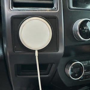 May include: A white wireless charging pad with a white cord attached to it. The charging pad is mounted in a black plastic housing. The charging pad is in a car dashboard.