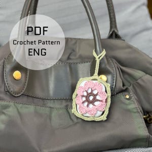 May include: A crocheted flower-shaped accessory in pink and brown, attached to a white square, hangs from a grey handbag. The accessory is framed by a green border and a green cord. The image includes the text "PDF Crochet Pattern ENG".