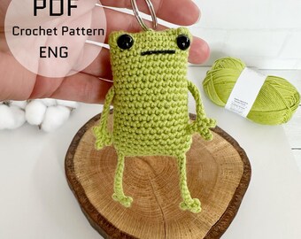 May include: A light green crocheted frog keychain with black eyes and a silver keyring. The frog is standing on a wooden slice. The text "PDF Crochet Pattern ENG" is in the upper left corner. A ball of green yarn is in the background.