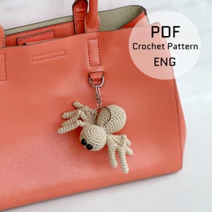May include: A crocheted spider keychain with black eyes and a beige body hangs from a coral-colored leather handbag. The keychain is attached with a silver-colored ring. Text on the image reads "PDF Crochet Pattern ENG".