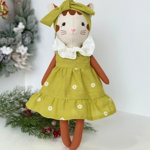 May include: A handmade plush cat doll wearing a green dress with white flowers and a green headband. The doll has brown fur and a white face with pink cheeks.