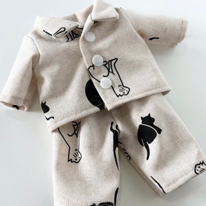 Puede incluir: Un conjunto de pijama de color crema con ilustraciones de gatos negros. La parte superior es una camisa de manga larga con cuello y cierre de botones. La parte inferior es un pantal&oacute;n de corte holgado.