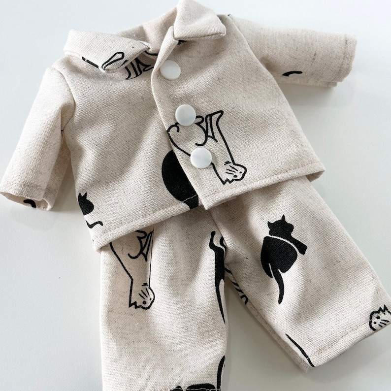 Puede incluir: Un conjunto de pijama de color crema con ilustraciones de gatos negros. La parte superior es una camisa de manga larga con cuello y cierre de botones. La parte inferior es un pantal&oacute;n de corte holgado.